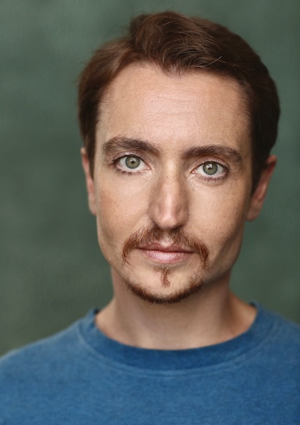 Headshot of white man looking at the camera and smiling. He has short brown hair and goatee and blue green eyes. He wears a blue T-shirt