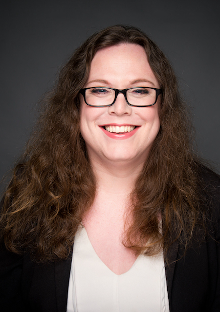 Headshot of white woman smiling and looking at the camera. She hash wavy brown hair and blue eyes. She wears black glasses, black jacket and white T-shirt.